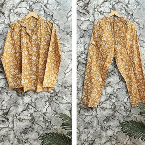 May include: A yellow and white floral print pajama set with long sleeves and pants. The set is hanging on a wooden hanger against a textured background.