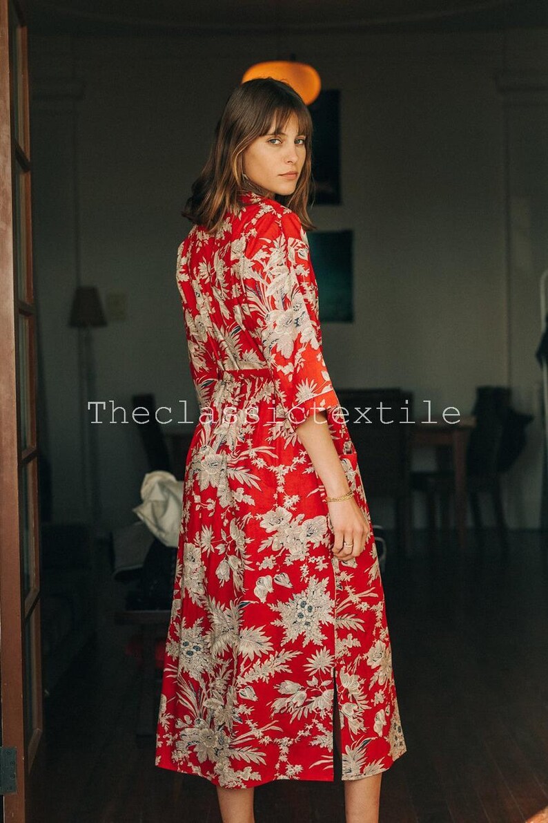 May include: A woman wearing a red and white floral midi dress with a tie waist. The dress has a side slit and is made of a lightweight fabric.