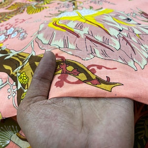 May include: Pink fabric with a floral print featuring yellow, brown, and red flowers. The fabric is being held by a hand.