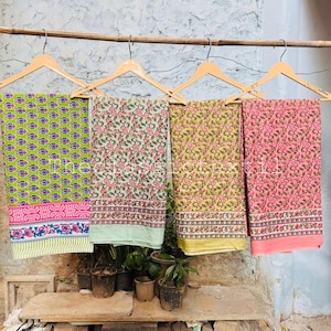 May include: Four colorful patterned fabrics hanging on wooden hangers. The fabrics feature floral and geometric designs in shades of green, pink, and yellow. Each fabric has a decorative border. The fabrics are displayed against a rustic background.