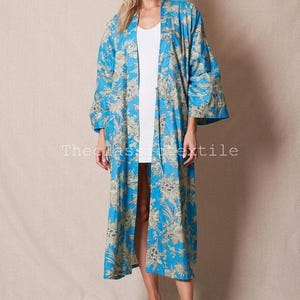 May include: A long, blue floral print kimono with a white background. The kimono has a wide, open collar and long sleeves. The model is wearing the kimono over a white vest top and sandals.