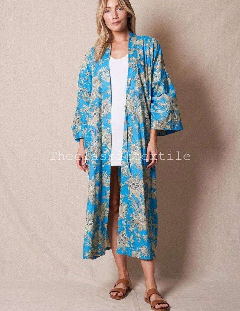 May include: A long, blue floral print kimono with a white background. The kimono has a wide, open collar and long sleeves. The model is wearing the kimono over a white vest top and sandals.