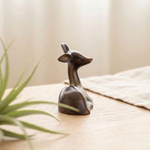 May include: A small, dark brown, smooth-surfaced figurine depicting the head and neck of a deer. The sculpture features a slender neck, pointed ears, and a gentle, forward-facing expression, crafted with a solid, minimalist form and a matte finish.