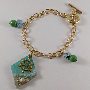 May include: A gold chain bracelet with a turquoise blue and white resin charm featuring a seashell and a gold ship's wheel. The bracelet also has two green beads and a toggle clasp.