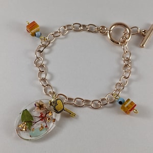 Pode incluir: Pulseira de corrente dourada com uma variedade de pingentes, incluindo um pingente de resina com flores prensadas e flocos dourados, um pequeno pingente de chave, um pingente de olho turco azul e um pingente listrado multicolorido.