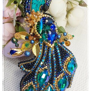 May include: A beaded peacock brooch with a blue and green iridescent design. The peacock's head is beaded with gold and black beads. The tail feathers are made of green and blue iridescent beads.