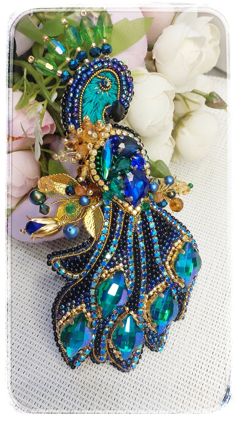 May include: A beaded peacock brooch with a blue and green iridescent design. The peacock's head is beaded with gold and black beads. The tail feathers are made of green and blue iridescent beads.