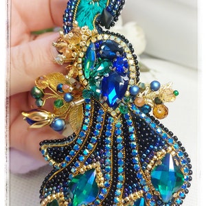 May include: A peacock brooch with blue, green, and gold accents. The peacock's tail feathers are made of iridescent green and blue rhinestones. The brooch is a beautiful and intricate piece of jewelry.
