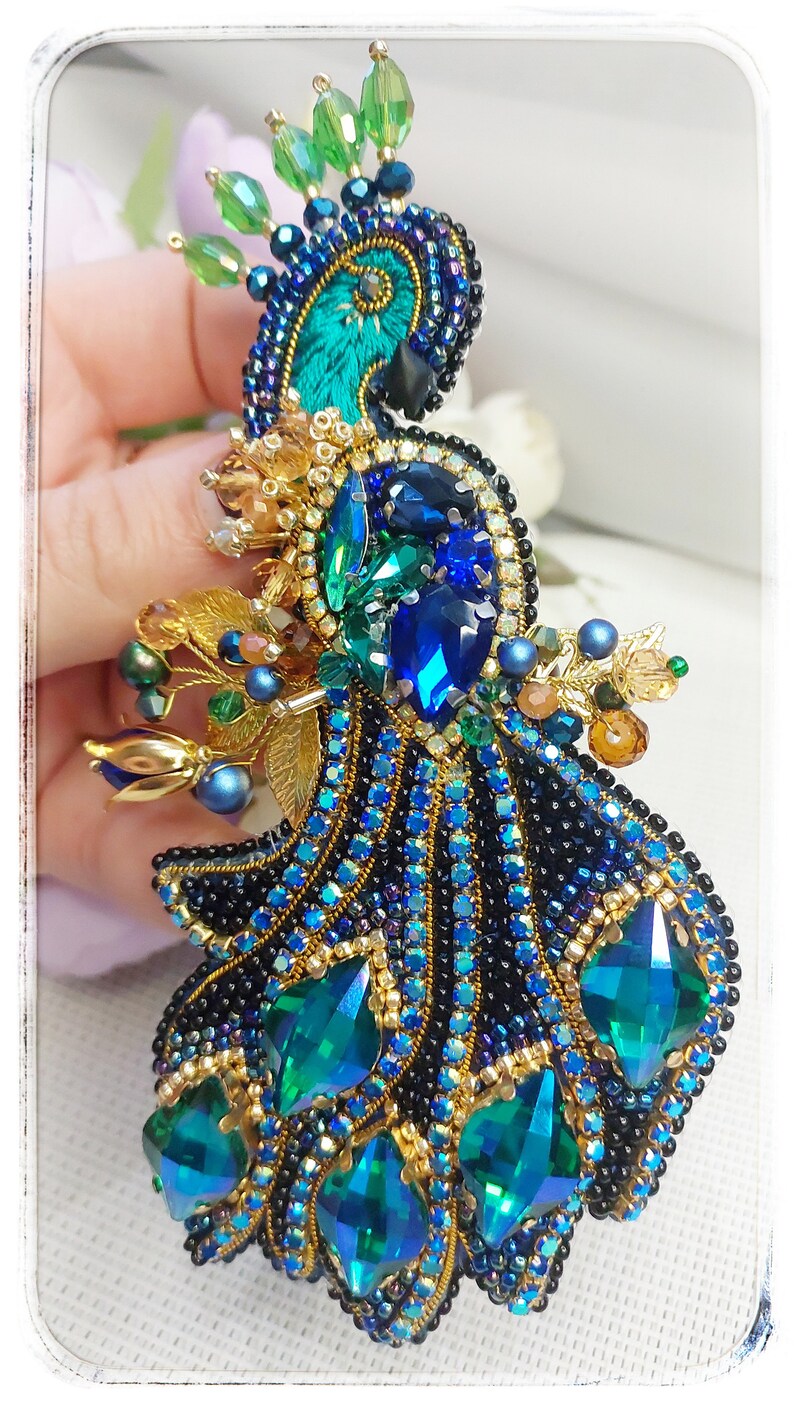 May include: A peacock brooch with blue, green, and gold accents. The peacock's tail feathers are made of iridescent green and blue rhinestones. The brooch is a beautiful and intricate piece of jewelry.
