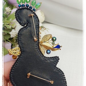 May include: A black leather brooch with gold and blue embellishments. The brooch is decorated with beads, sequins, and a gold leaf design. It has a pin back for attaching to clothing.
