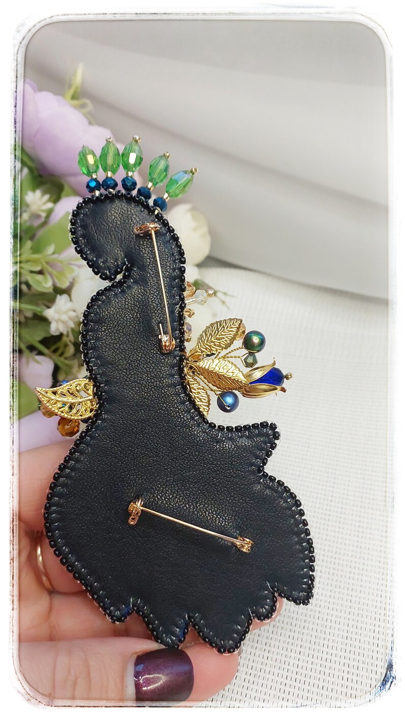 May include: A black leather brooch with gold and blue embellishments. The brooch is decorated with beads, sequins, and a gold leaf design. It has a pin back for attaching to clothing.
