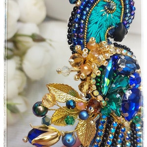 May include: A peacock-inspired brooch with blue, green, and gold beads and crystals. The brooch features a detailed peacock feather design with a gold trim.