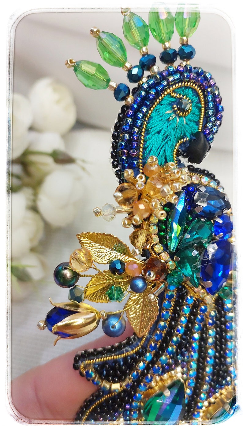 May include: A peacock-inspired brooch with blue, green, and gold beads and crystals. The brooch features a detailed peacock feather design with a gold trim.