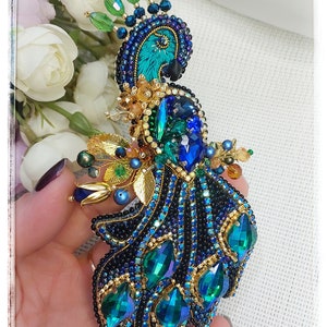 May include: A peacock brooch made of blue, green, and gold beads. The peacock's tail feathers are spread out and have a iridescent sheen.