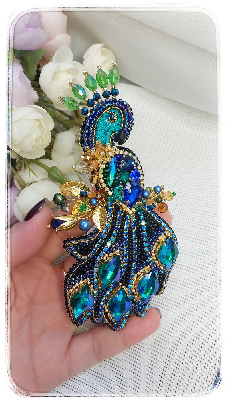 May include: A peacock brooch made of blue, green, and gold beads. The peacock's tail feathers are spread out and have a iridescent sheen.