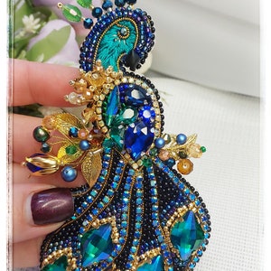 May include: A beaded peacock brooch with a blue and green color scheme. The peacock's tail feathers are made of green and blue rhinestones. The peacock's head is made of black and blue beads. The brooch is adorned with gold accents.