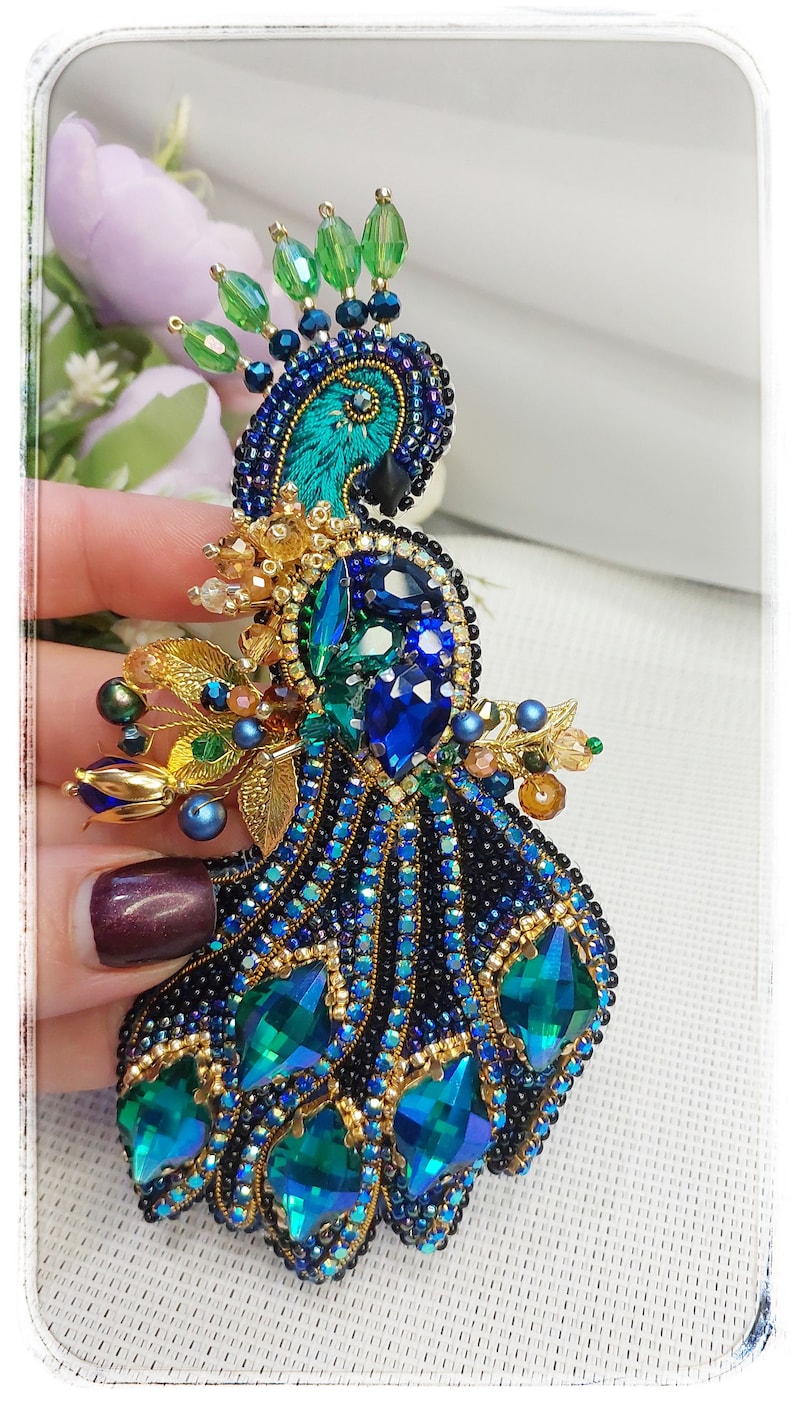 May include: A beaded peacock brooch with a blue and green color scheme. The peacock's tail feathers are made of green and blue rhinestones. The peacock's head is made of black and blue beads. The brooch is adorned with gold accents.