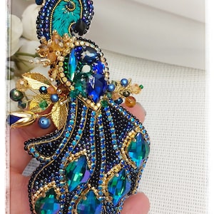 May include: A beaded peacock brooch with blue, green, and gold accents. The peacock's tail feathers are made of iridescent green and blue beads.