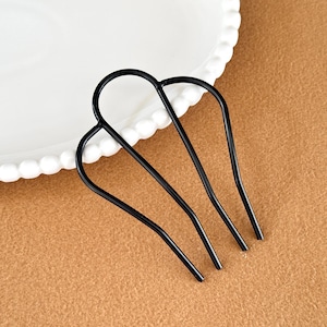 Puede incluir: Un peine de pelo de metal negro con tres púas, apoyado sobre una superficie marrón. El peine tiene una parte superior curvada y está junto a un plato blanco con un borde decorativo. Es un accesorio para el cabello.