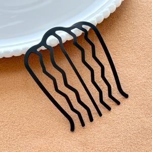 May include: A black metal hair comb with a wavy design. The comb has six curved teeth and a curved top. The comb is on a peach-colored surface, near a white plate with a scalloped edge. Hair accessory.