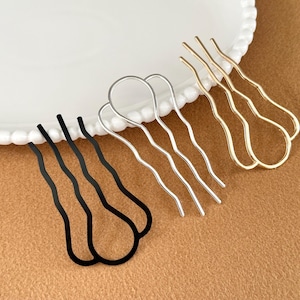 May include: Three hair pins in black, silver, and gold colors. The pins have a wavy design and are arranged on a white plate with a scalloped edge. The background is a solid tan color.