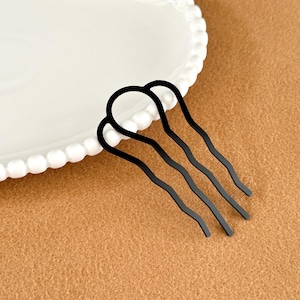 May include: A black hair comb with a wavy design. The comb is made of metal and has three prongs. It is lying on a tan surface next to a white plate with a decorative edge. This hair accessory is suitable for various hairstyles.