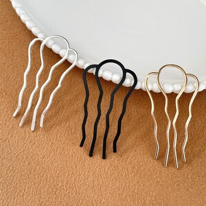 May include: Three metal hair combs in silver, black, and gold. Each comb has a wavy design with three prongs. The combs are arranged on a white plate with a beaded edge, set against a brown surface.