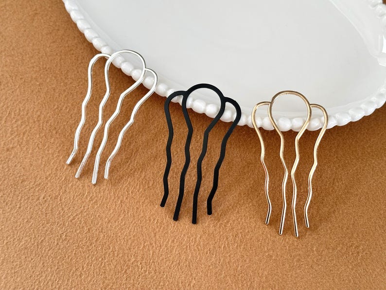 May include: Three metal hair combs in silver, black, and gold. Each comb has a wavy design with three prongs. The combs are arranged on a white plate with a beaded edge, set against a brown surface.