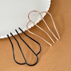 May include: Two hair pins. One is black, the other is gold-colored. Both have a U-shaped design with three prongs. The hair accessories are displayed on a white plate with a decorative edge and a brown surface.