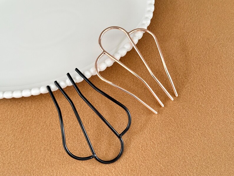 May include: Two hair pins. One is black, the other is gold-colored. Both have a U-shaped design with three prongs. The hair accessories are displayed on a white plate with a decorative edge and a brown surface.