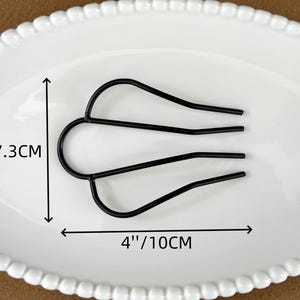 May include: Black metal hair comb, 4 inches (10cm) wide and 2.9 inches (7.3cm) tall, resting on a white oval plate. The comb has a U-shaped top with three tines extending downward. The plate has a beaded edge.
