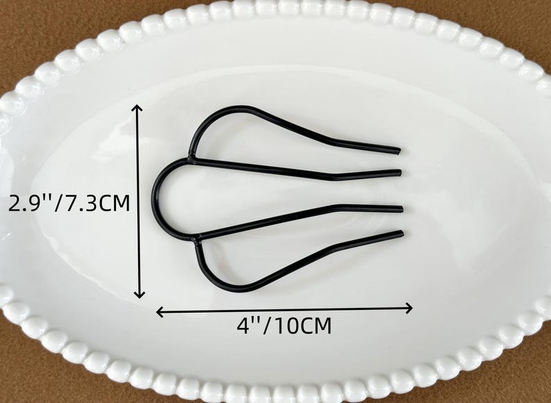 May include: Black metal hair comb, 4 inches (10cm) wide and 2.9 inches (7.3cm) tall, resting on a white oval plate. The comb has a U-shaped top with three tines extending downward. The plate has a beaded edge.