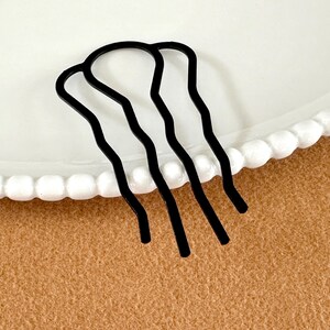 May include: A black metal hair comb with three wavy prongs. The hair accessory is on a white plate with a scalloped edge, set against a tan background. This hair comb is designed to secure and style hair.