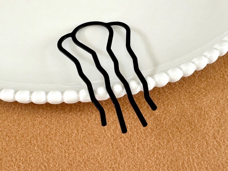 May include: A black metal hair comb with three wavy prongs. The hair accessory is on a white plate with a scalloped edge, set against a tan background. This hair comb is designed to secure and style hair.