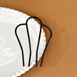 May include: Three black hair pins arranged on a white, scalloped-edge plate. The pins have a simple, curved design. The background is a solid, warm brown color.