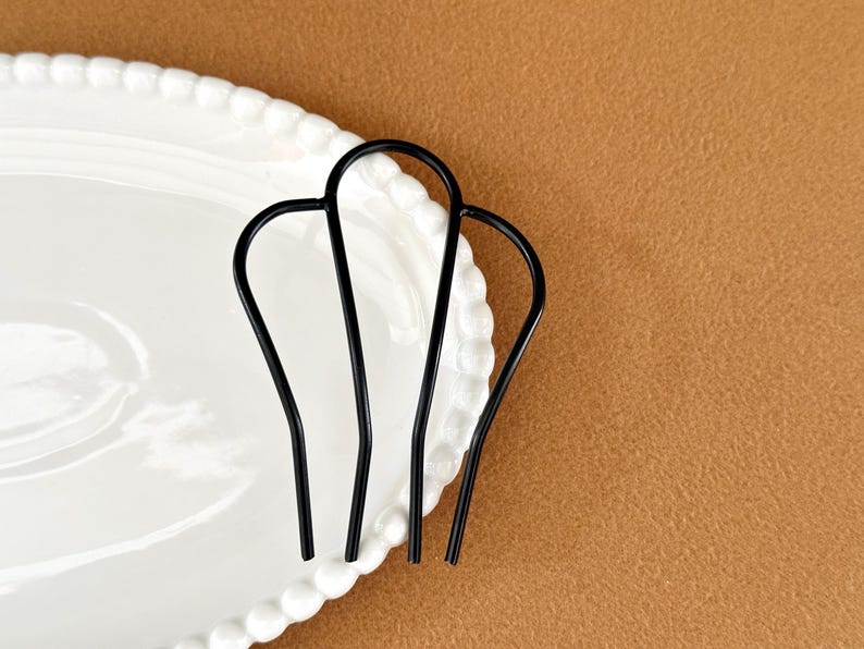 May include: Three black hair pins arranged on a white, scalloped-edge plate. The pins have a simple, curved design. The background is a solid, warm brown color.