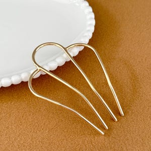 May include: A gold-colored hair comb with a curved design. The comb has four tines and is positioned on a brown surface next to a white plate with a beaded edge. The comb is likely used for styling hair.