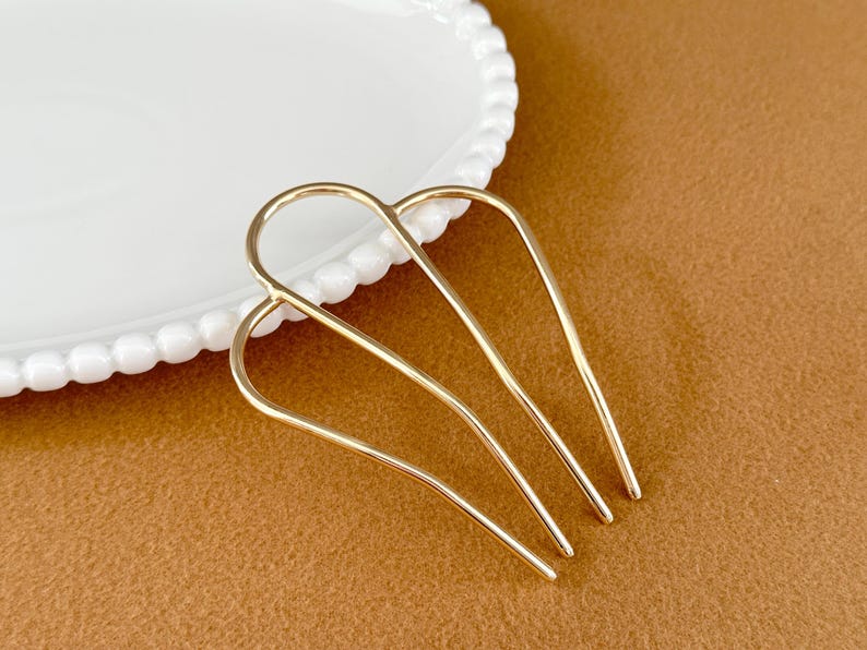 May include: A gold-colored hair comb with a curved design. The comb has four tines and is positioned on a brown surface next to a white plate with a beaded edge. The comb is likely used for styling hair.