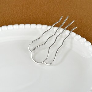 May include: A pair of silver hair pins with a wavy design, resting on a white oval plate with a beaded edge. The background is a solid brown color.