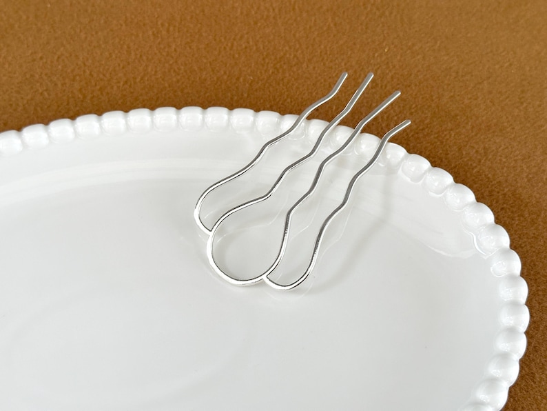 May include: A pair of silver hair pins with a wavy design, resting on a white oval plate with a beaded edge. The background is a solid brown color.