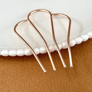 May include: Three rose gold hair pins arranged on a white plate with a beaded edge, set against a brown background. The hair pins are U-shaped with a smooth, metallic finish. These hair accessories are designed for styling and securing hair.