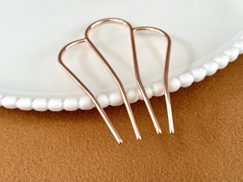 May include: Three rose gold hair pins arranged on a white plate with a beaded edge, set against a brown background. The hair pins are U-shaped with a smooth, metallic finish. These hair accessories are designed for styling and securing hair.