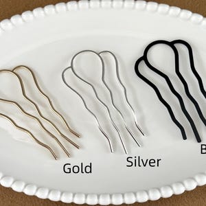 May include: Three metal hair pins in gold, silver, and black, arranged on a white, scalloped-edge plate. The pins have a wavy design and are labeled with their respective colors.