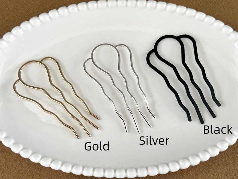 May include: Three metal hair pins in gold, silver, and black, arranged on a white, scalloped-edge plate. The pins have a wavy design and are labeled with their respective colors.