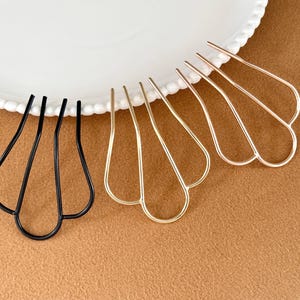 May include: Hair accessories: A collection of hair pins in black and gold tones. The pins have a unique, openwork design with a petal-like shape. They are arranged on a white plate with a scalloped edge, set against a brown background.