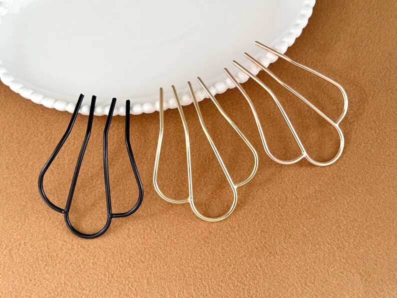 May include: Hair accessories: A collection of hair pins in black and gold tones. The pins have a unique, openwork design with a petal-like shape. They are arranged on a white plate with a scalloped edge, set against a brown background.