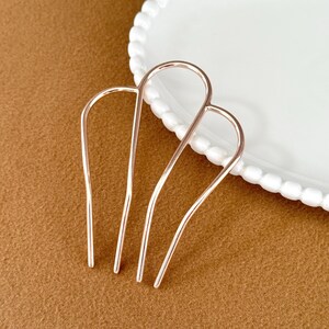 May include: Two rose gold hair pins are displayed on a brown surface next to a white plate with a beaded edge. The hair accessories are U-shaped with three prongs each, designed for securing hair in place.