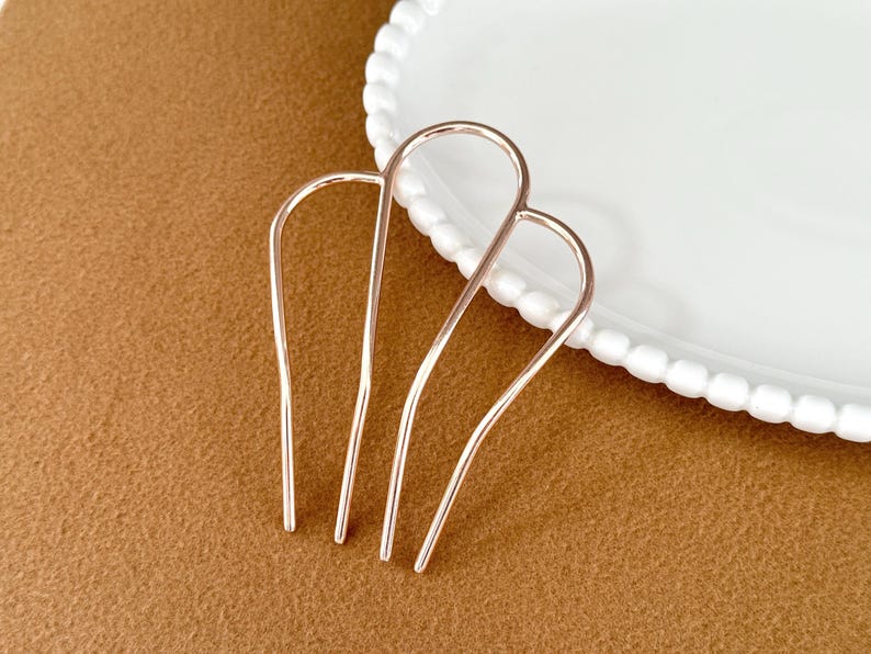 May include: Two rose gold hair pins are displayed on a brown surface next to a white plate with a beaded edge. The hair accessories are U-shaped with three prongs each, designed for securing hair in place.