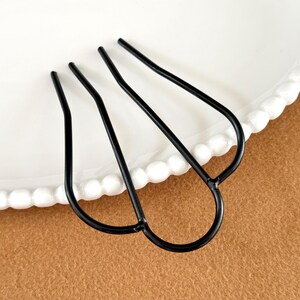 May include: A black metal hair fork with three prongs, resting on a white plate with a beaded edge. The hair accessory is set against a brown background, showcasing its elegant design.
