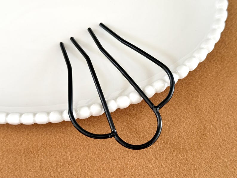 May include: A black metal hair fork with three prongs, resting on a white plate with a beaded edge. The hair accessory is set against a brown background, showcasing its elegant design.
